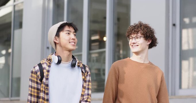 Upon entering frame, male friends walking with headphones outside building noticing glass facade