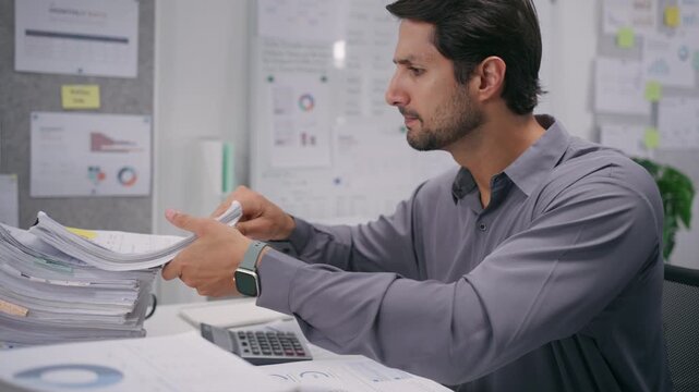 Latin man reviews financial accounting documents and tax paperwork, checking each report and file in an office.