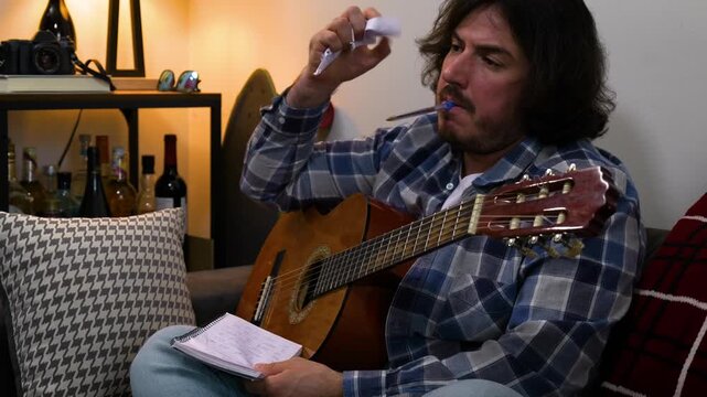 A man is sitting on his sofa with his guitar, gets frustrated writing music on his notebook, tears the paper and starts again.