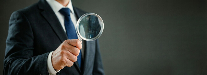 Businessman in suit holding magnifying glass, concept of financial scanner on dark background.