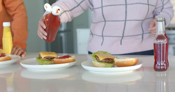 Friends at kitchen counter, flipping cap, woman squeezing ketchup onto bun and man grabbing mustard