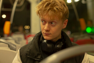 young man sitting on bus headphones around neck, gazing toward window, empty carriage bathed in warm lights, layered jacket and backpack visible, thoughtful expression, latenight urban commute,