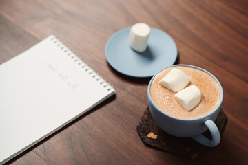 Hot chocolate in a blue cup with marshmallow and notepad on walnut table