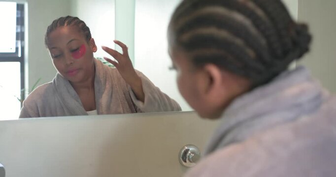 Glancing in bathroom mirror at sink African American woman applying under-eye patches for hydration