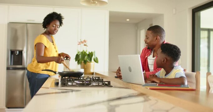 African American family in kitchen mother stirring pan, talking father pausing typing, son nodding