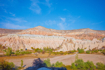 A vibrant landscape photograph of Cappadocia, Turkey, showcasing its famous fairy chimney formations, layered pastel cliffs, and warm desert terrain under a bright, clear blue sky