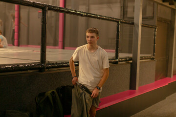 Young man leaning on railing, waiting between rehearsals in dim industrial corridor, wearing white shirt and jeans, backpack on floor, pink neon accents, relaxed posture and contemplative expression,