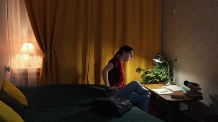 Young Caucasian woman student sits at desk at home studying in evening room with warm light preparing for university exams and focusing on education process. Wide shot.