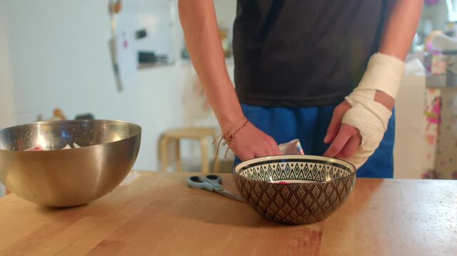 bandaged hands arranging small items into patterned bowl on wooden worktop, kitchen backdrop with supplies,