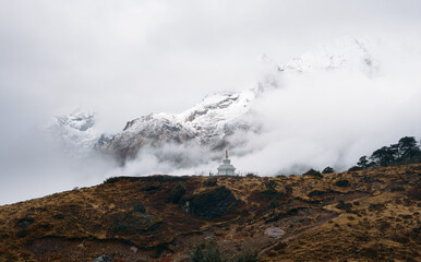 Clouds cover snowy peaks near a stupa in the mountains during an overcast day while mist drifts around the landscape