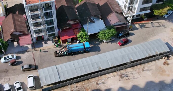 Aerial view of a blue garbage truck with sanitation workers collects waste along a sunlit urban residential street with apartments and houses.