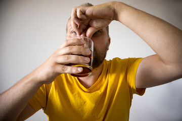 A man in a yellow T-shirt pinches his nose while sipping a dark drink from a glass, capturing a candid reaction to an unpleasant taste or odor.