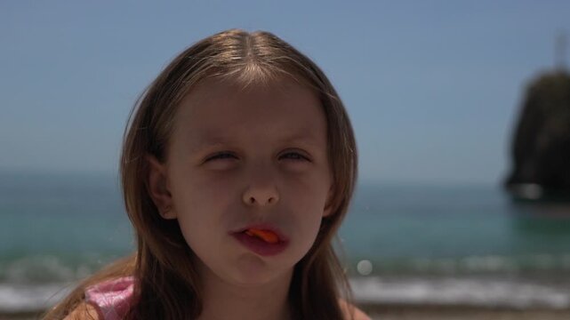Girl, beach, lollipop, young girl chews orange candy on a sunny seaside vacation.