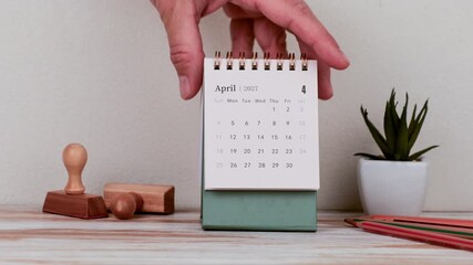 April 2027 Calendar Page Displayed On A Desk As A Hand Adjusts The Monthly Planner