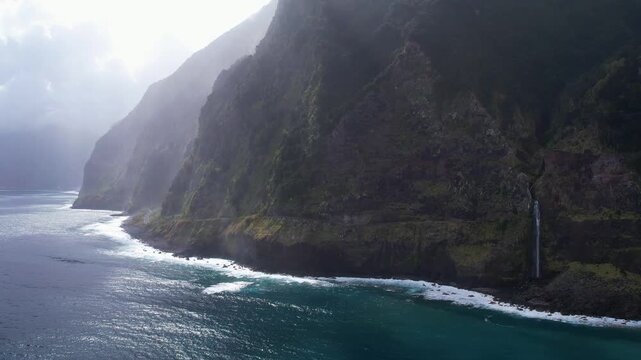 Aerial Madeira coastal scene with a waterfall Veu da Noiva plunging down steep cliffs into the ocean. The rugged landscape merges with the dynamic waves, creating a breathtaking natural spectacle