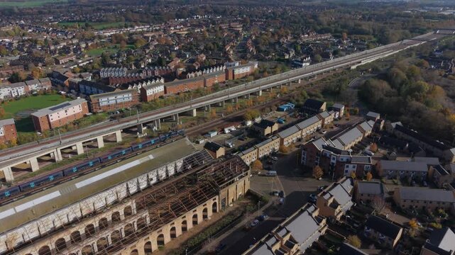 Train leaving Ashford town in Kent, UK. Aerial view following the train journey. Flying above to old warehouse and the town. Local train driving next to a fast speed international railway tracks.