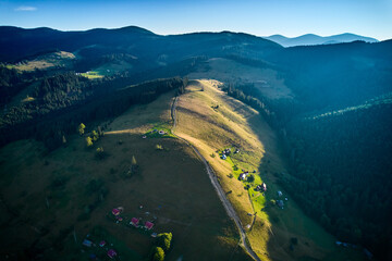 Obraz premium Aerial view of picturesque valley with rolling hills, lush forests, and scattered houses. Sunlight bathes landscape in warm glow, highlighting vibrant greens and creating serene atmosphere.