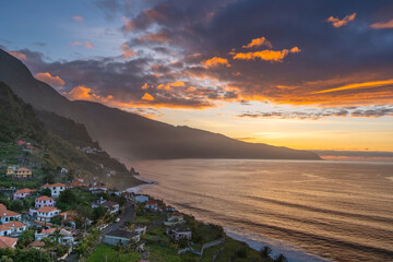 Dramatic aerial view of Madeira's coastline at sunset, showcasing vibrant oranges and blues. The serene ocean, lush mountains, and charming village create a captivating scene