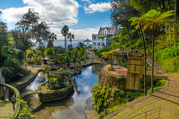 A serene garden scene in Madeira Monte Palace tropical garden in Funchal with lush greenery, a tranquil stream, and traditional architecture in the background. Ideal for nature lovers and tourists.