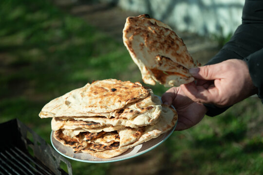 Hand holding a piece of golden-brown grilled flatbread over a stack of fresh bread on a plate with a sunny outdoor BBQ setup.