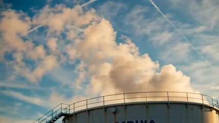 Industrial storage tank releasing steam against a vibrant sky with clouds and contrails