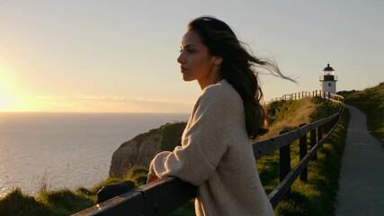 A woman in a cozy sweater gazing at the sunset beside a lighthouse, with ocean waves crashing below
