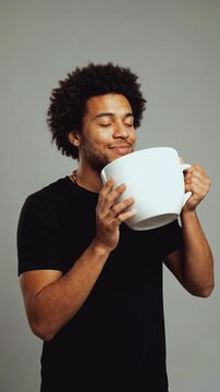 Vertical video of a young man with an afro holding a comically large white mug. Happy male smelling the aroma of coffee or tea with a smile. Humorous concept of caffeine addiction