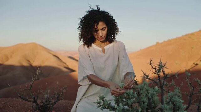 A young woman in a light dress tending to a plant in a desert landscape with rolling hills at sunset