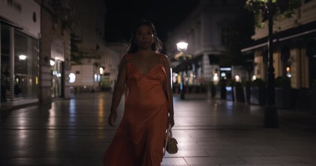 Asian woman in elegant orange dress walking confidently down an illuminated city street at night, embodying urban sophistication and late-night adventure
