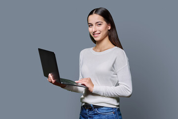 Portrait of smiling teenage girl holding laptop on gray studio background