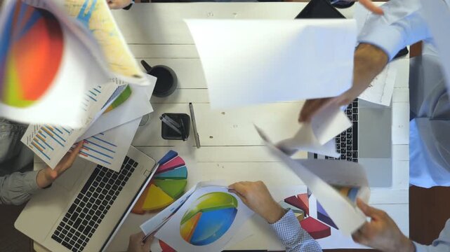 Hands of business people celebrating achievement throwing up documents with graphs. Workers rejoicing success clapping their hands. Creative team working at modern office. Slow mo Top view