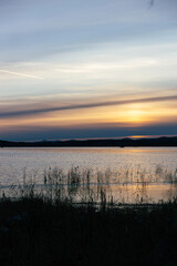 Obraz premium Serene sunset over tranquil lake, with gentle ripples reflecting warm hues of orange and purple, surrounded by distant hills and soft clouds, creating a peaceful natural landscape