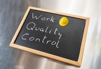 Small chalkboard with the text Work Quality Control and yellow hard hat. Concepts of quality control, and standards.Blackboard on a metallic background