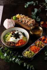 Enjoy a delightful spread of Korean cuisine with bulgogi, fresh vegetables, sushi rolls, and savory soup on a dark wooden table, surrounded by lush greenery