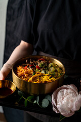 A warm bowl of colorful noodles piled high with fresh vegetables and savory toppings rests on a dark tray. A small dish of sauce complements this inviting meal, creating a feast for the senses