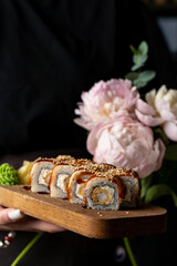 Artfully arranged sushi rolls showcase fresh ingredients and vibrant colors. A hand holds the wooden platter adorned with beautiful peonies, creating a warm and inviting atmosphere