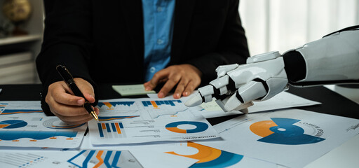 Business professional analyzing financial charts with robotic arm assisting in data interpretation and decision-making at a modern office setting. Noogenesis