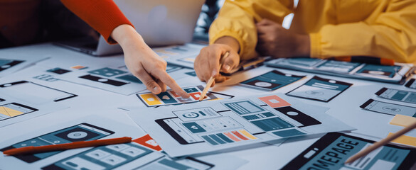 Close-up of a collaborative workspace featuring hands selecting user interface mockups from a table. Laptops and digital tools create an engaging environment for creative professionals. SACTR