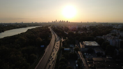 Obraz premium Smooth drone footage flying toward the Warsaw city skyline during a warm summer sunset. The video captures the Vistula River, extensive green areas, residential neighborhoods, and a major highway fil