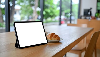 Digital tablet with blank white screen mockup resting on a wooden cafe table next to a croissant and coffee cup, with a blurred modern background.