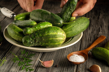 Fresh cucumbers are placed on a plate while a hand picks one up. A spoon with salt and garlic lies nearby on a wooden table in a kitchen. The setting is bright and natural