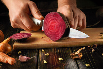 A person is slicing a vibrant beetroot on a wooden cutting board. Nearby, there are carrots, garlic, onions, and spices creating a rustic kitchen atmosphere. The focus is on fresh ingredients