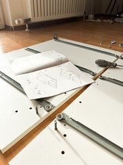 White furniture assembly process at home. Instruction manual, screwdriver, screws, and furniture parts on a wooden laminate floor