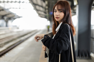 Young Asian businesswoman waiting at train station, checking smartwatch while commuting. Concept of urban lifestyle, time management, professional mobility and smart technology in daily routine.