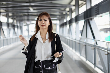 Confident young Asian businesswoman walking on a modern skywalk, holding tablet and stylus. Concept of mobile work, tech savvy professionals and urban lifestyle.