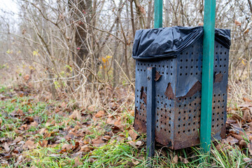 Fototapeta premium A trash bin stands near a trail in a forest, surrounded by dry leaves and bare trees under an overcast sky