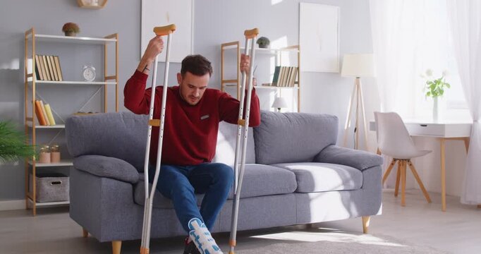 Man is about to get up from sofa and is using crutches for support due to leg injury. Caucasian young man with broken limb who wears ankle bandage and uses crutches to walk. Injury concept. 4K video.