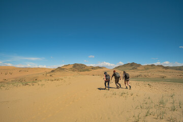 Three adventurers with backpacks trek across the sand dunes of the Gobi Desert in Mongolia. Rare green shrubs dot the landscape, with rocky mountains under a vast, clear blue sky in the distance.