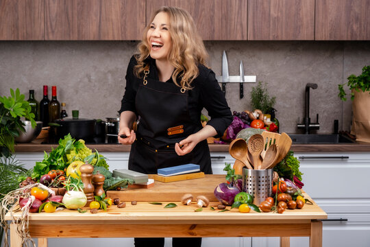Laughing blonde woman in black chef apron stands at wooden kitchen table with fresh vegetables and knife sharpening stones in bright home kitchen. Cooking, culinary lifestyle, healthy food concept.