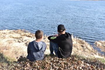 Two boys sitting on a grassy riverbank and looking at the calm water on a sunny day.